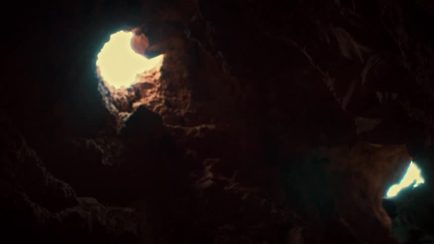 A low angle view shows a woman in silhouette climbing up the rocky walls to escape a dark cave through an opening to the daylight