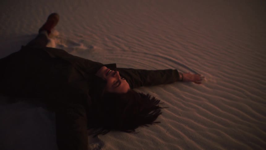 A joyful woman lies on a rippled sand dune at dusk, smiling as she makes a sand angel