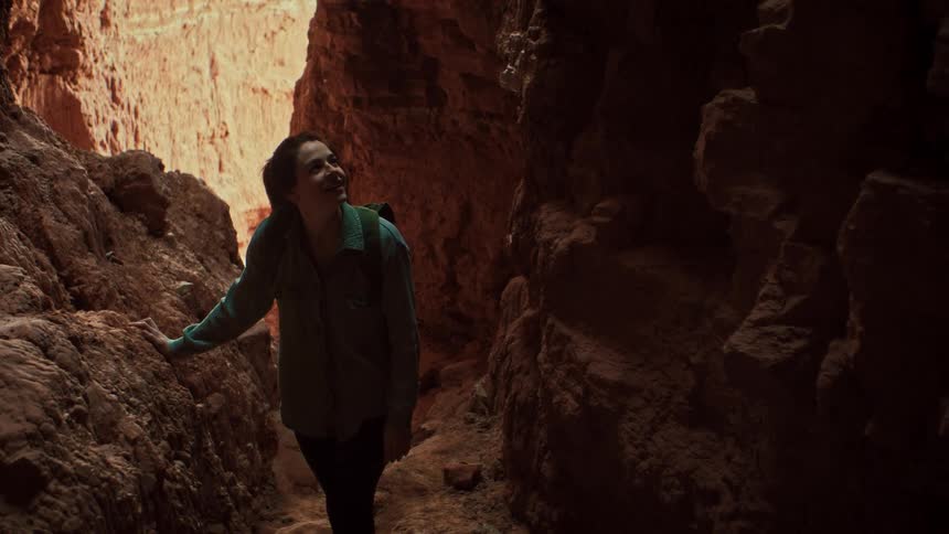A female hiker with a backpack stands in awe inside a narrow red rock slot canyon, looking up towards the light