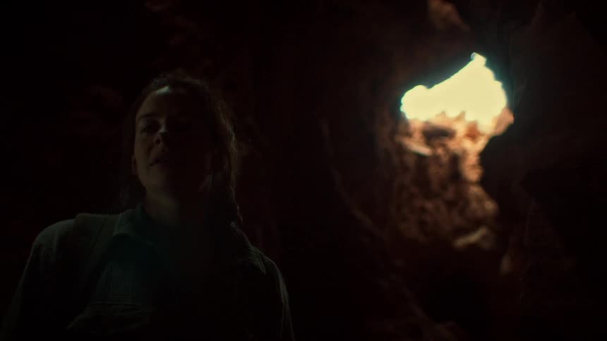 An adventurous woman pauses to look at a beam of light from an opening above while exploring a dark cave, then turns and continues into the darkness