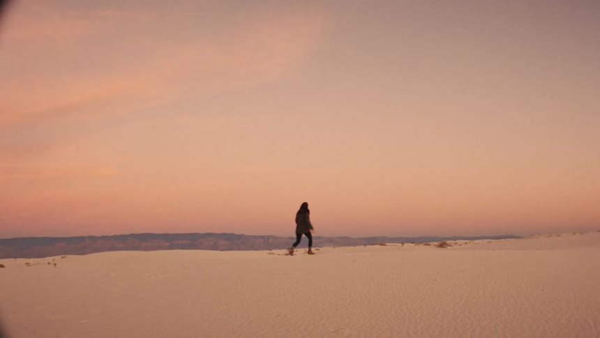 A carefree woman walks in slow motion through a vast white sand desert, playfully kicking up sand against a beautiful sunset sky