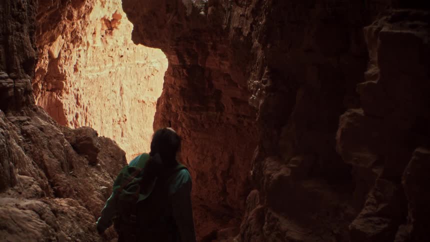 A young woman with a backpack gazes up in wonder as she hikes through a narrow, dark slot canyon in slow motion