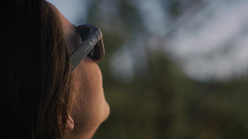 A happy woman wearing sunglasses smiles as she looks up towards the sky during a serene golden hour sunset