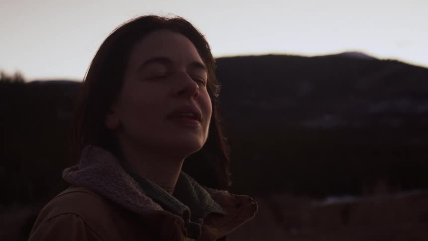 A serene woman closes her eyes, basking in the last light of the day against a mountain silhouette