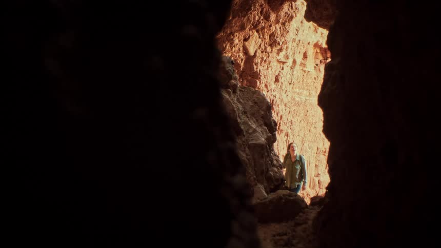 A female hiker with a backpack explores a sunlit red rock canyon, viewed from inside a dark cave