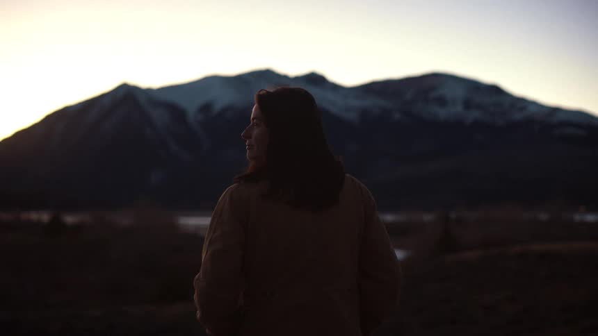 A woman in silhouette looks out at a majestic snow-capped mountain range at sunset in slow motion