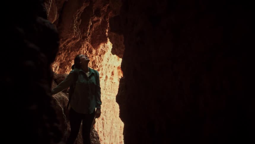 An awe-struck female hiker with a backpack stands inside a dark cave, looking up at the sunlit opening above