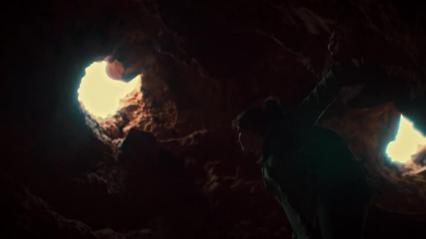A silhouetted explorer climbs through a dark rock cave, looking up at the bright sunlight shining through holes in the ceiling