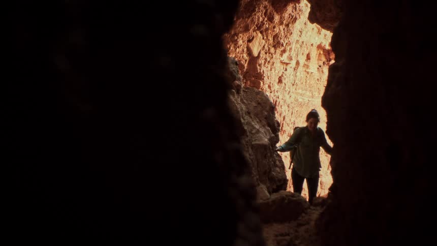 A female hiker in slow motion carefully explores a narrow, sunlit passage from within a dark desert cave