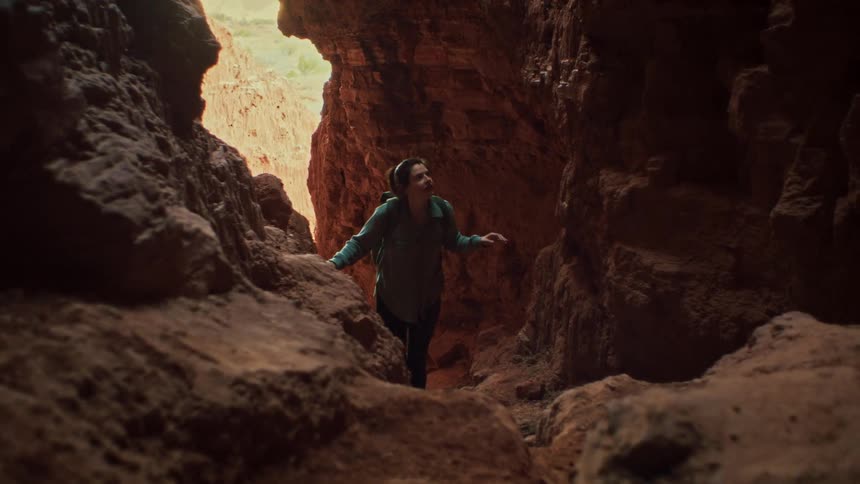 An adventurous woman slowly hikes through a dark, narrow red rock canyon, gazing up in awe at the majestic rock formations