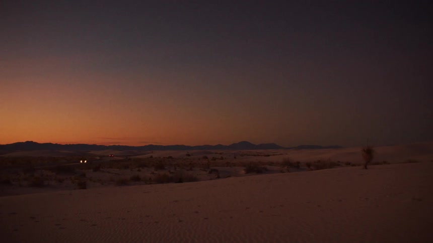 Distant car headlights illuminate a highway running through the vast white sand desert at twilight