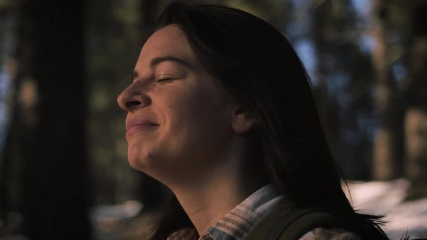 A serene woman closes her eyes and smiles, enjoying a moment of peace while hiking in a sunlit winter forest