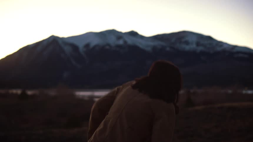 A solo female adventurer takes off her backpack to admire the serene view of a snow-capped mountain range at twilight