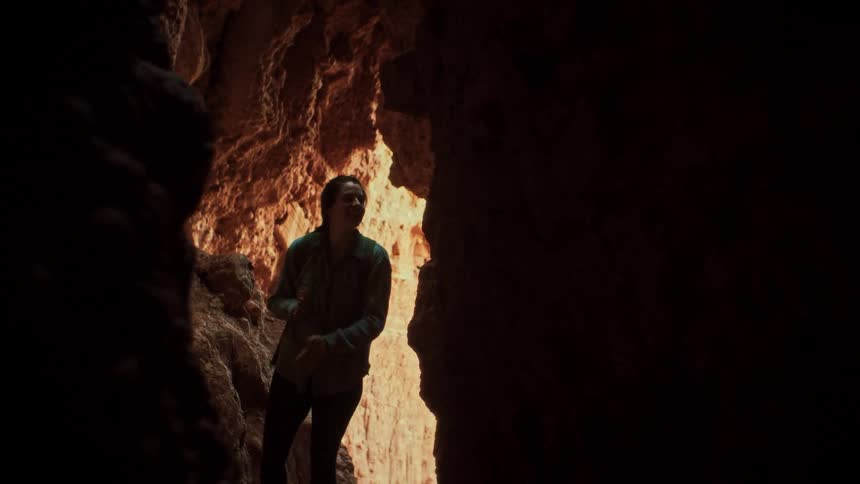 An adventurous woman explores a dark and narrow cave, illuminated by a sliver of light as she marvels at the rock formations