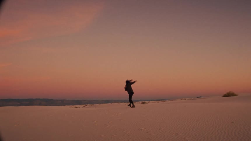 A woman joyfully runs and jumps across the white sand dunes in slow motion as the sun sets in the distance