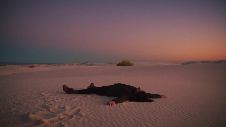 A lone figure lies peacefully on the white sand of a desert during a beautiful sunset