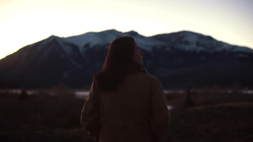 A woman wearing a coat stands peacefully while looking out at a snow-capped mountain range during a serene sunset