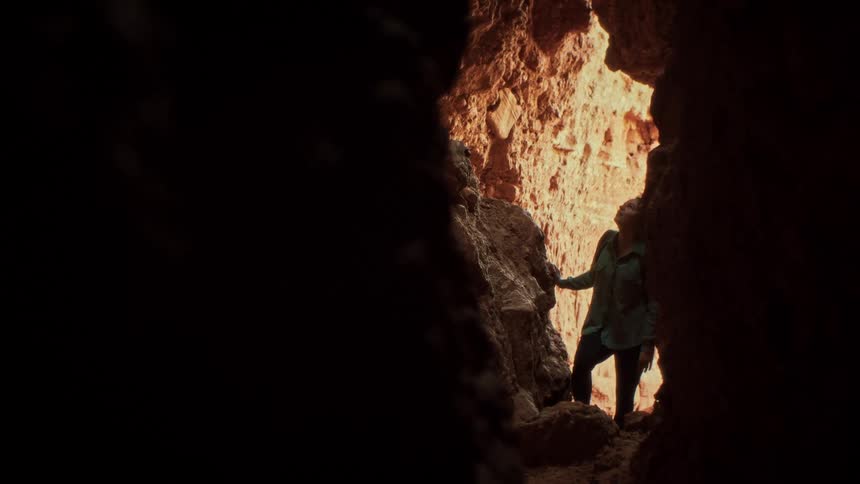 A woman cautiously explores a glowing red rock canyon from the darkness of a cave in slow motion
