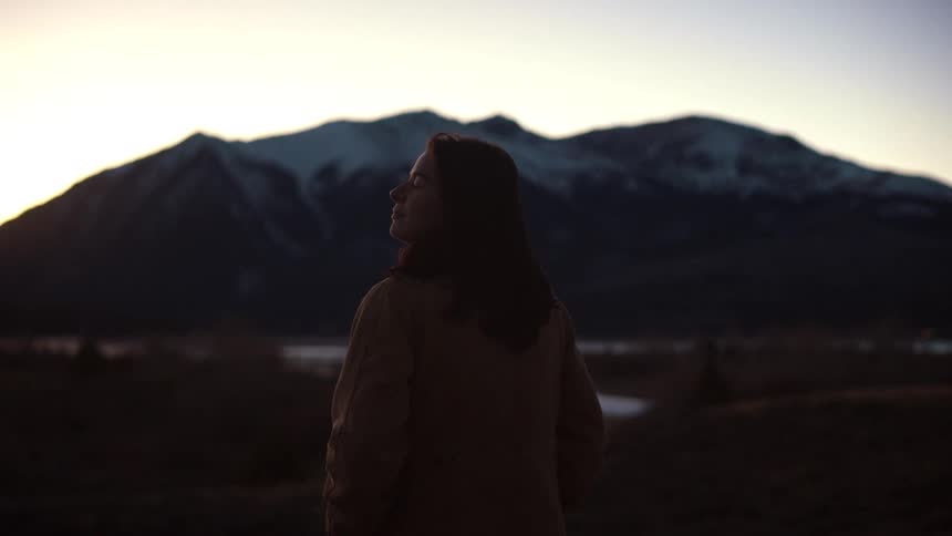 A calm woman in a jacket takes a deep breath and smiles while enjoying the mountain view at sunset