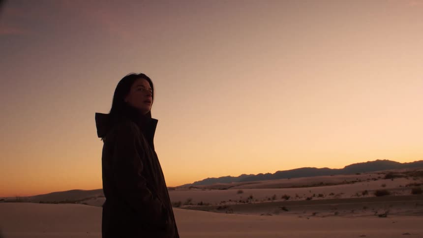 A woman in a hooded jacket stands in a vast sand dune desert at sunset, peacefully watching the sky over distant mountains