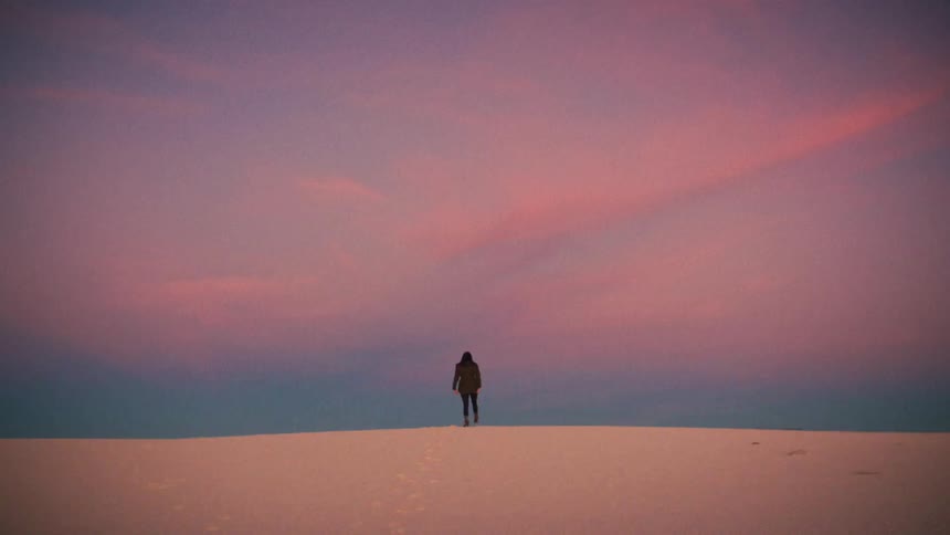 A lone figure rises from a vast white sand dune and walks in solitude, stretching their arms in freedom under a vibrant pink and purple sunset sky