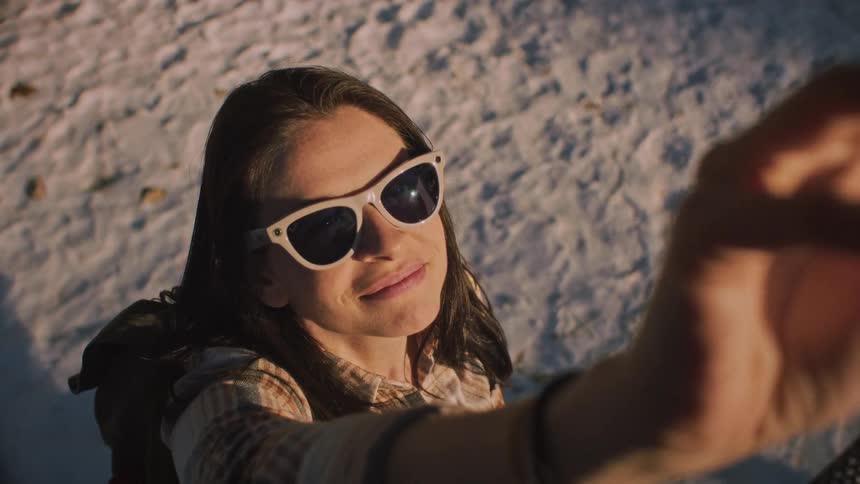 High angle slow-motion shot of a smiling woman in sunglasses shielding her eyes from the bright sun on a snowy day