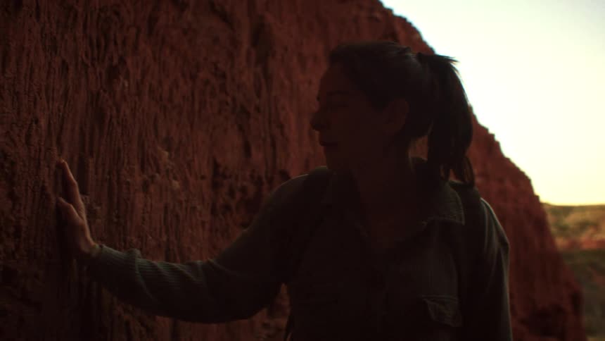 A silhouetted woman smiles in awe at the view from a red rock canyon alcove before touching the textured wall in slow motion