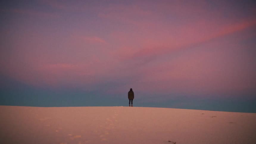 A solitary figure stands atop a white sand dune, watching the vibrant pink and purple sunset sky