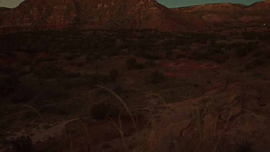 A lone hiker traverses a dusty trail in a shadowy red rock canyon as night begins to fall