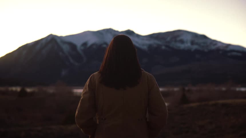 A woman stands still from behind, taking in the view of a dramatic, snow-capped mountain range at dusk