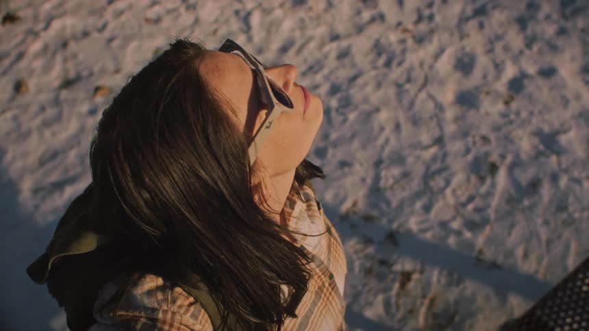 A smiling woman with a backpack looks up to soak in the sun's warmth on a snowy day before turning to continue her hike