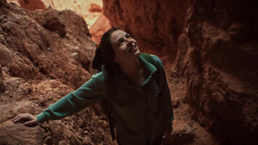 A young female hiker smiles in awe as she looks up at the light filtering into a beautiful red rock slot canyon