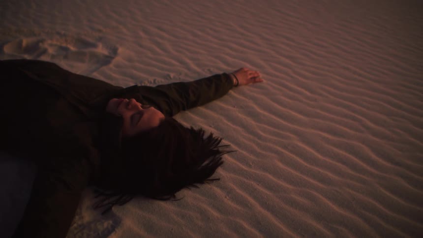 A serene person with long dark hair lies peacefully on the rippled sand dunes at sunset