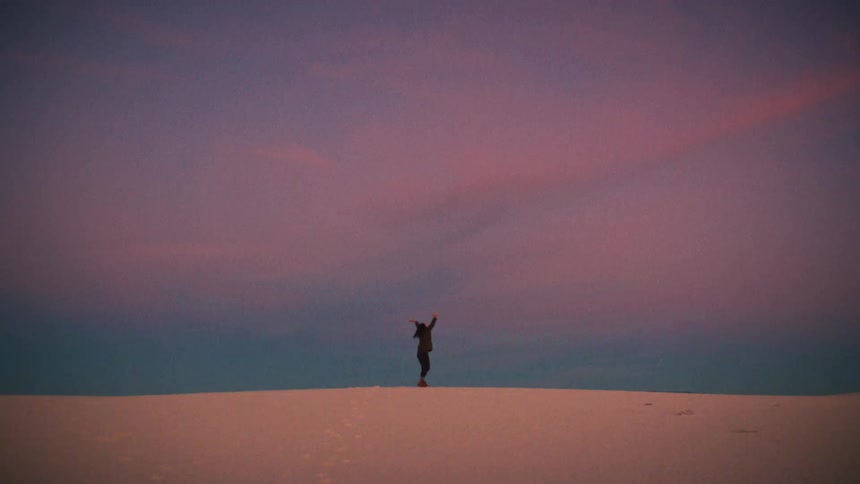 A lone figure raises their arms in celebration on top of a vast sand dune under a stunning pastel sunset