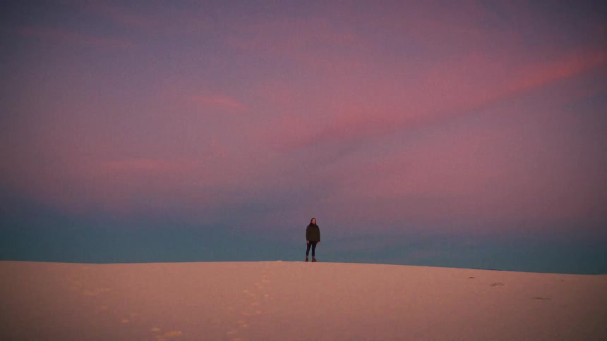 A lone figure walks along the ridge of a vast white sand dune under a beautiful pink and purple sunset sky