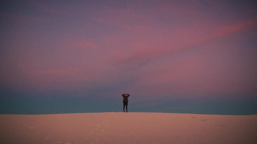 A lone person triumphantly raises their arms while standing atop a white sand dune under a beautiful pink and purple twilight sky