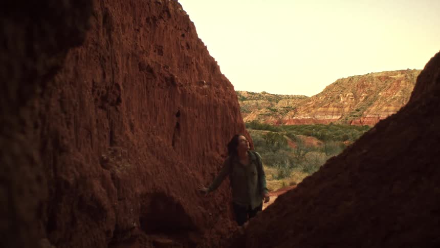 A woman hiker with a backpack slowly explores a narrow red rock canyon, looking up in awe at the natural formations
