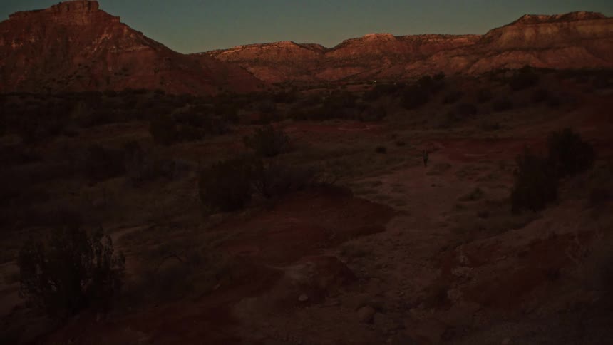 A lone hiker treks through a vast, quiet desert canyon at twilight as the last rays of sunlight illuminate the distant red rock mesas