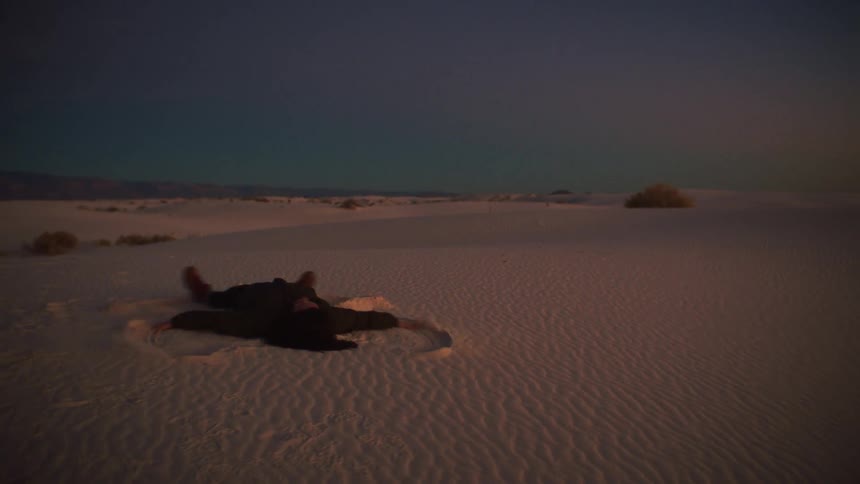 A woman makes a sand angel on a vast white sand dune at dusk before getting up and running with joy