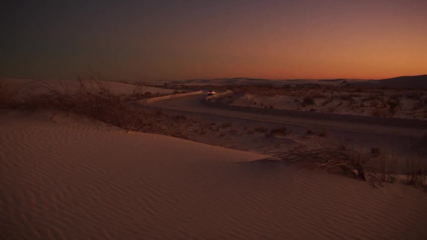 A white sedan drives along a winding road through a serene white sand desert landscape at sunset