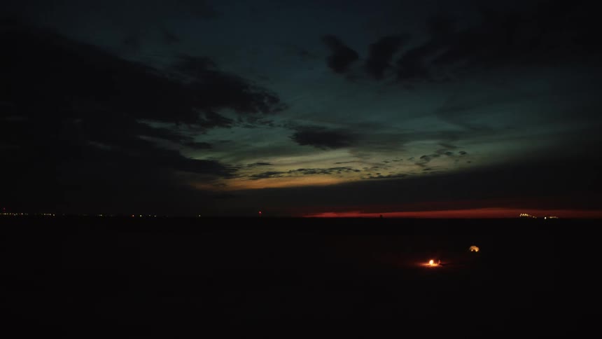 A small campfire is lit in a vast, dark landscape under the last colors of sunset
