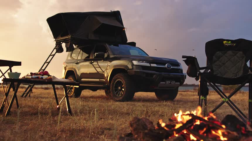 Rugged off-road vehicle with a rooftop tent parked at a serene lakeside campsite with a crackling campfire during a golden sunset
