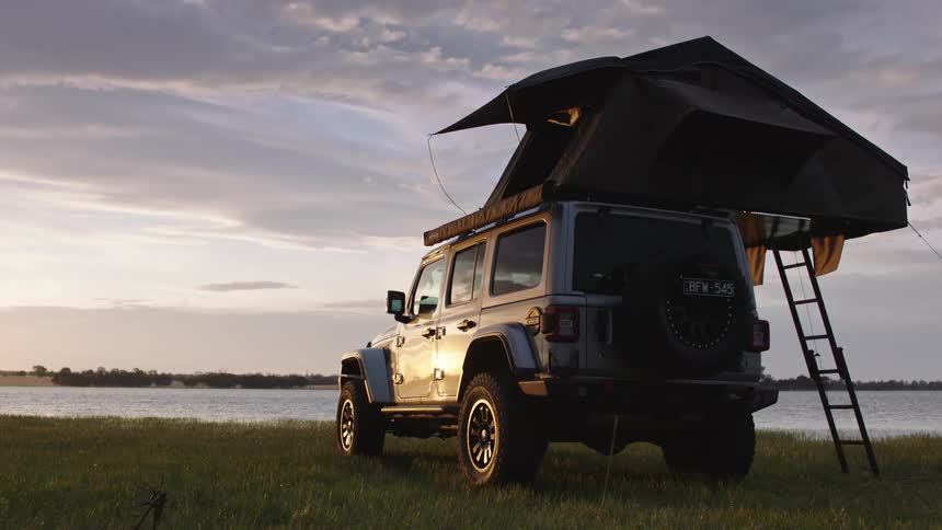 A rugged Jeep with a pop-up rooftop tent is parked in a grassy field by a serene lake during a beautiful sunset