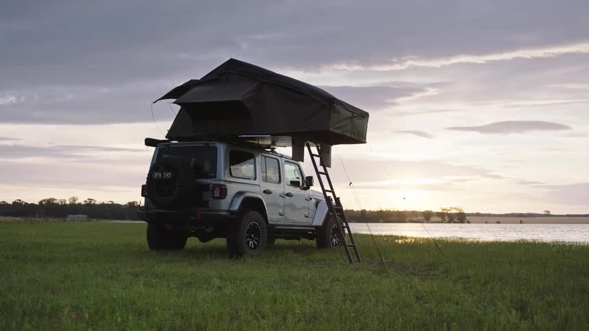 A modern Jeep with a pop-up rooftop tent is set up for camping in a grassy field beside a lake at sunset