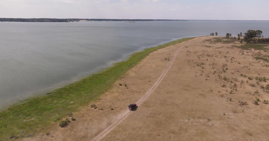 High-angle aerial shot of a red car driving on a dirt road alongside a vast lake, kicking up a trail of dust