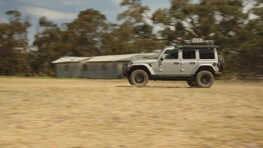 A silver Jeep equipped for overlanding drives off-road through a dry, grassy field in the outback
