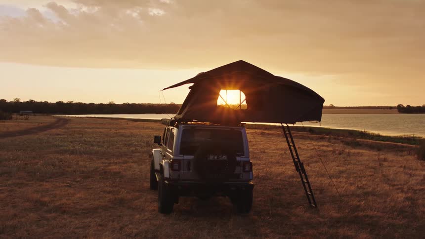 A white off-road vehicle with a rooftop tent is parked by a tranquil lake at sunset, with the golden light shining through the window