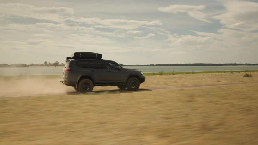 Slow motion tracking shot of a dark 4x4 driving along a dusty lakeside track, creating a plume of dust in its wake