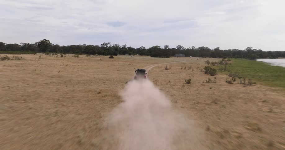 A drone chases a red off-road SUV as it speeds down a dusty dirt road through a vast, dry field next to a lake