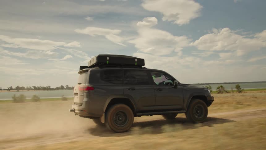 An off-road SUV equipped with a rooftop tent kicks up dust as it drives along a scenic lakeside dirt road in slow motion